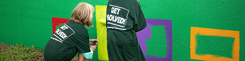 Two young girls paint a mural over the top of a graffitied wall.